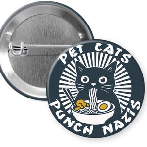 May include: A round, navy blue button with a black cat eating ramen. The text reads "PET CATS PUNCH NAZIS" in a white, stylized font. The button has a metal pin back.
