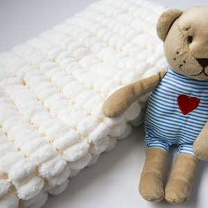 May include: A white crocheted blanket with a brown teddy bear wearing a blue and white striped shirt with a red heart on it.