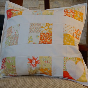 May include: Square cushion with a patchwork design. The cushion features white fabric with sections of floral patterns in orange, yellow, and green. A white heart applique is on the lower right corner. The cushion is placed on a wooden chair.