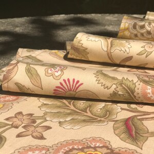 May include: Folded fabric with a floral design. The cream-coloured fabric features large flowers in red, brown, and yellow, with green leaves and stems. The pattern creates a classic, vintage look. The fabric is folded, showing the detailed design.