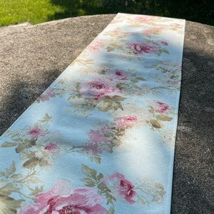 May include: A light blue floral table runner with a pattern of pink and cream roses. The rectangular runner, likely fabric, is designed to decorate a table. The floral design adds a touch of elegance to any dining table or surface. The runner measures approximately 150 cm in length.