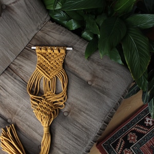 May include: A mustard yellow macrame wall hanging with a geometric pattern and a long tassel. The hanging is displayed on a gray textured surface.