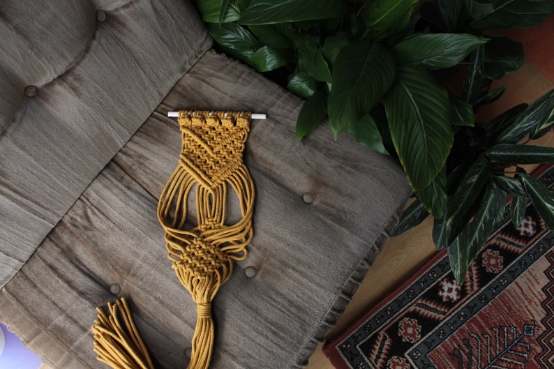 May include: A mustard yellow macrame wall hanging with a geometric pattern and a long tassel. The hanging is displayed on a gray textured surface.