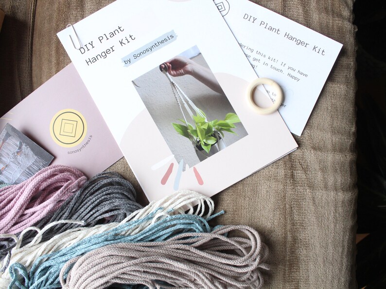 May include: DIY plant hanger kit with instructions, a wooden ring, and four spools of macrame cord in pink, gray, white, and blue.