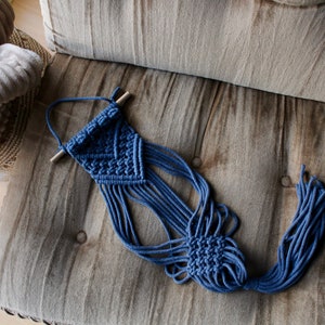 May include: A blue macrame wall hanging with a wooden dowel at the top. The hanging has a geometric pattern and long fringe.