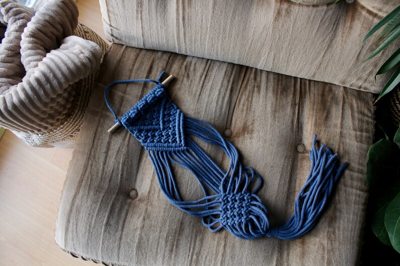 May include: A blue macrame wall hanging with a wooden dowel at the top. The hanging has a geometric pattern and long fringe.