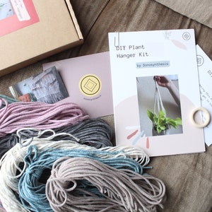 May include: DIY plant hanger kit with instructions, four spools of macrame cord in different colors, and a wooden ring. The kit is on a brown fabric surface.