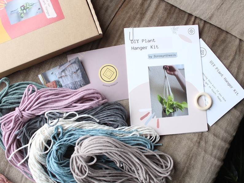 May include: DIY plant hanger kit with instructions, four spools of macrame cord in different colors, and a wooden ring. The kit is on a brown fabric surface.