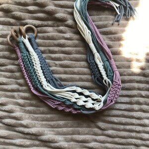 May include: A set of four macrame plant hangers with wooden rings. The hangers are made with a combination of white, gray, and pink cotton cord.