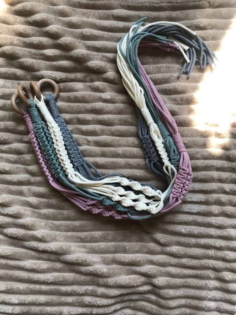 May include: A set of four macrame plant hangers with wooden rings. The hangers are made with a combination of white, gray, and pink cotton cord.