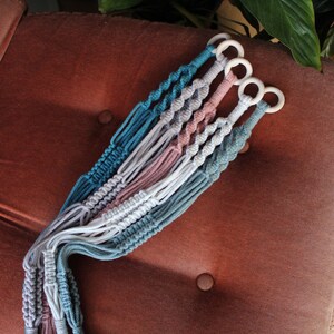 May include: Four macrame plant hangers in teal, white, pink, and gray with wooden rings at the top.