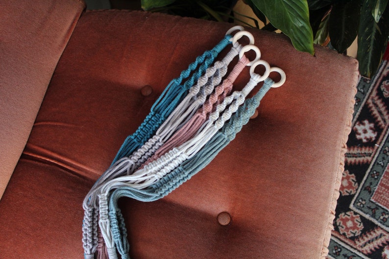 May include: Four macrame plant hangers in teal, white, pink, and gray with wooden rings at the top.