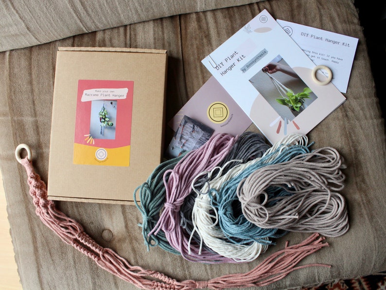 May include: DIY plant hanger kit with instructions, a wooden ring, and four skeins of yarn in pink, gray, blue, and beige.
