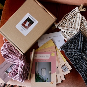 May include: A DIY wall hanging kit with two macrame wall hangings, one in white and one in grey, with pink yarn and instructions. The kit includes a wooden dowel, yarn, and instructions.