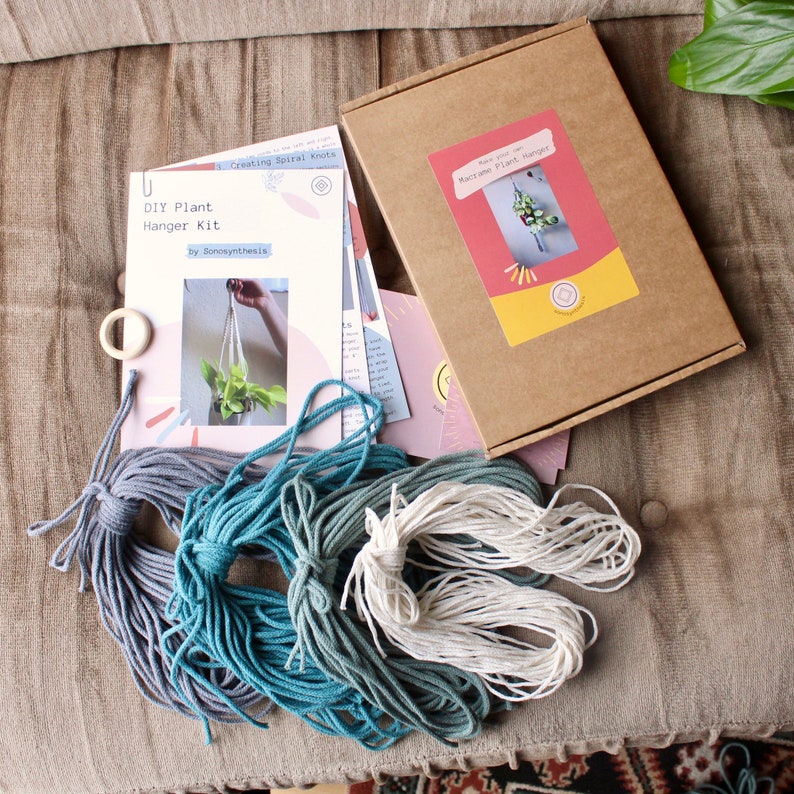 May include: DIY macrame plant hanger kit with instructions, three skeins of yarn in shades of blue and white, and a wooden ring. The kit includes instructions for creating a spiral knot macrame plant hanger.