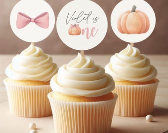 May include: Three cupcakes with white frosting and decorative toppers. The toppers feature a pink bow, the text "Violet is one" with a pumpkin, and a pumpkin illustration. The cupcakes are on a light brown surface.