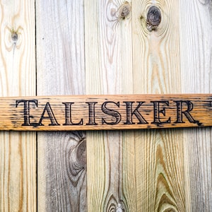 Può includere: Un cartello di legno con la parola "TALISKER" bruciata sulla superficie. Il cartello è fatto di legno marrone chiaro con una venatura naturale.