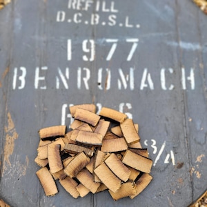 Op de afbeelding: Een close-up van een stapel kleine, rechthoekige stukjes hout. Het hout is lichtbruin van kleur en heeft een ruwe textuur. Het hout is in een willekeurig patroon gerangschikt. Het hout ligt op een grijs oppervlak. De tekst "REFILL D.C.B.S.L. 1977 BENROMACH" is zichtbaar op het oppervlak.