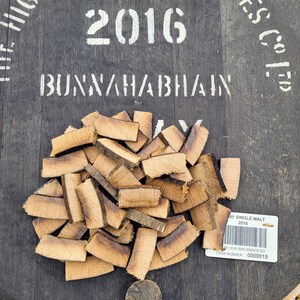 May include: A close-up of a pile of wooden barrel staves on a dark wooden surface. The staves are a light brown color and have a slightly charred appearance. The surface has the text "2016 BUNNAHABHAIN" and "ES COED" printed on it. A white label with the text "AY SINGLE MALT 2016" and a barcode is visible in the lower right corner. The label also includes the text "MT 2016 0000 0000019 GO" and "CASK NUMBER 0000019".
