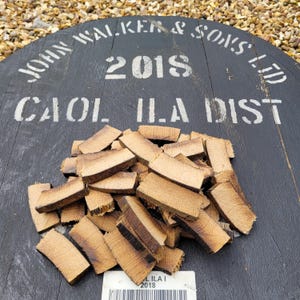 Op de afbeelding: Een close-up van een stapel bruine houtsnippers op een zwart houten oppervlak. Het oppervlak heeft witte letters met de tekst "John Walker & Sons Ltd Caol Ila Dist 2018".