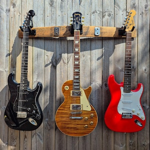 Peut inclure: Trois guitares électriques accrochées à un mur en bois. Les guitares sont noires, marron et rouges. Les guitares sont montées sur un support en bois. Le mur est fait de planches de bois.