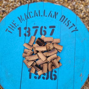 Op de afbeelding: Een blauwe houten vatentop met de tekst "THE MACALLAN DISTY" en de nummers "13767" en "1936". Een stapel kleine, bruine, rechthoekige stukjes hout bevindt zich in het midden van de vatentop.