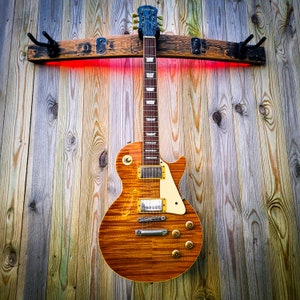 May include: An electric guitar with a sunburst finish, mounted on a wooden rack with red LED lighting. The guitar features a cream pickguard and gold hardware. The wall is made of weathered wooden planks.
