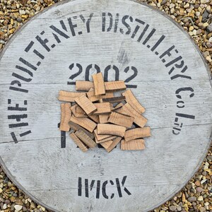 Op de afbeelding: Een close-up van een stapel kleine, lichtbruine houtsnippers op een wit houten oppervlak. Het oppervlak heeft zwarte letters met de tekst "The Pulterney Distillery Co. Ltd. 2002 Wick".
