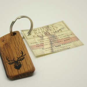 Könnte beinhalten: Ein Holzschlüsselanhänger mit einem Hirsch-Design, hergestellt aus einem alten Whiskyfass. Der Schlüsselanhänger ist an einem silbernen Ring befestigt. Das Etikett lautet "Whisky Barrel Keyrings" und "Barrel Craft Studio".