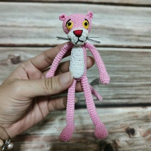 May include: Pink crocheted plush toy of the Pink Panther cartoon character. The toy has a white belly and black whiskers.