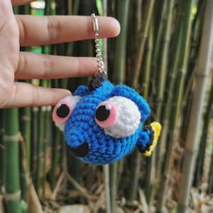 May include: A blue and white crocheted fish keychain with pink eyes and a yellow fin. The keychain is hanging from a silver chain.