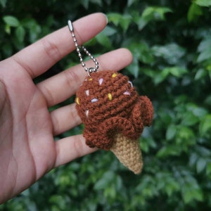 May include: A brown crocheted ice cream cone keychain with white and yellow sprinkles. The keychain is attached to a silver chain.