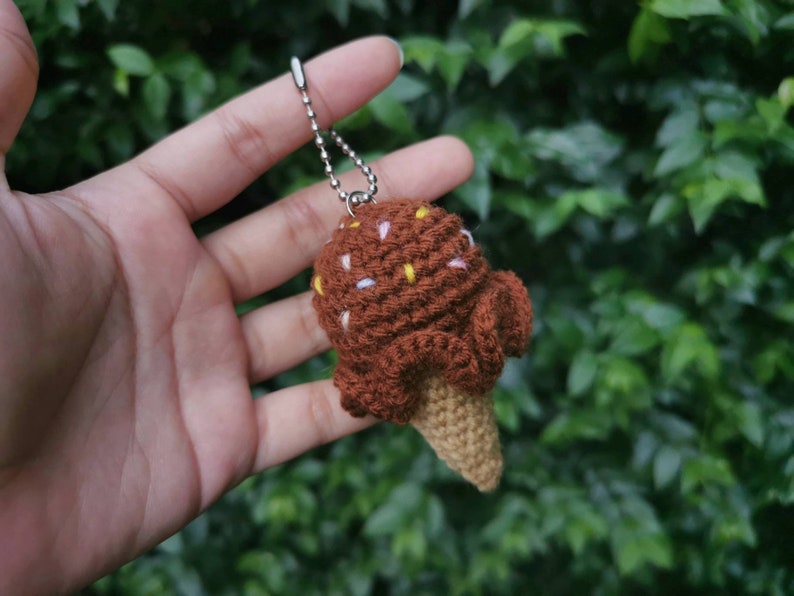 May include: A brown crocheted ice cream cone keychain with white and yellow sprinkles. The keychain is attached to a silver chain.