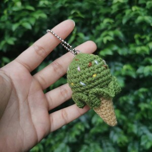 May include: A green crocheted ice cream cone keychain with white and brown sprinkles. The keychain is hanging from a silver chain.