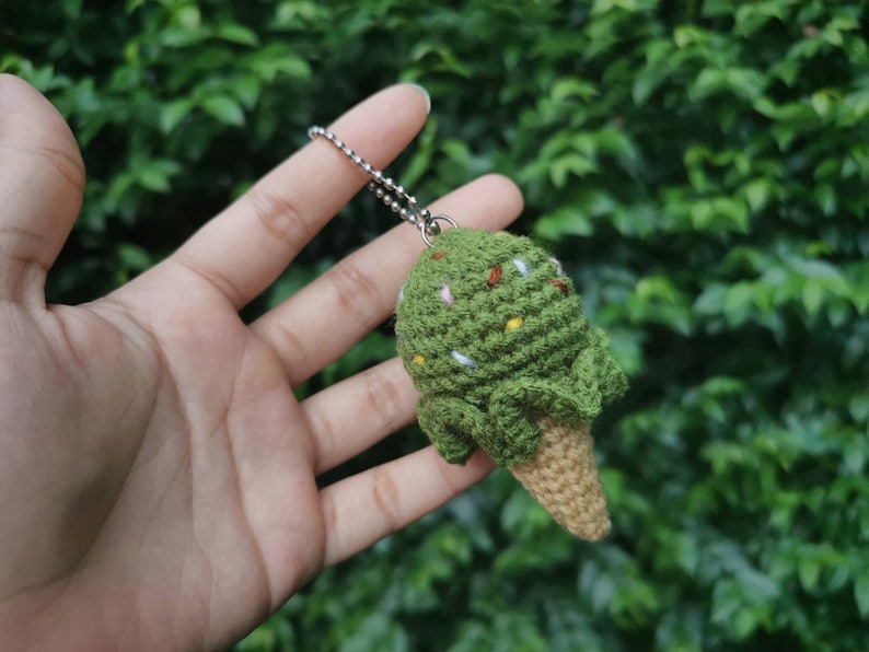 May include: A green crocheted ice cream cone keychain with white and brown sprinkles. The keychain is hanging from a silver chain.