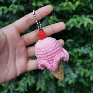 May include: A pink crocheted ice cream cone keychain with a red cherry on top. The keychain is held in a hand against a green leafy background.