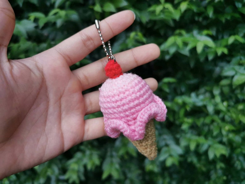 May include: A pink crocheted ice cream cone keychain with a red cherry on top. The keychain is held in a hand against a green leafy background.