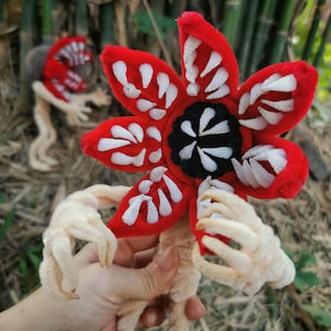 May include: A plush Demogorgon flower creature with a red, petal-like head featuring white embroidered teeth. The center is black with white accents. It has beige, textured limbs and is held by a hand. Another similar creature is in the background.