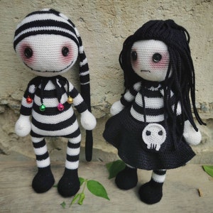 May include: Two crocheted dolls with black and white striped clothing. The doll on the left has a black and white striped hat and bells around its neck. The doll on the right has long black hair and a black skirt with a skull design.