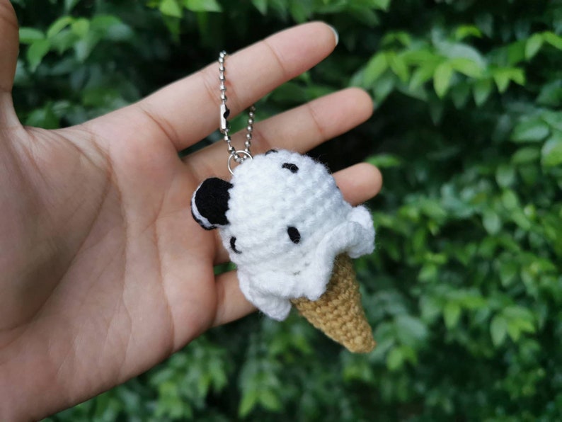 May include: A white crocheted ice cream cone keychain with black chocolate chips. The keychain is attached to a silver chain.