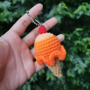 May include: A crocheted ice cream cone keychain with a red cherry on top. The cone is orange and brown and has a silver chain attached.
