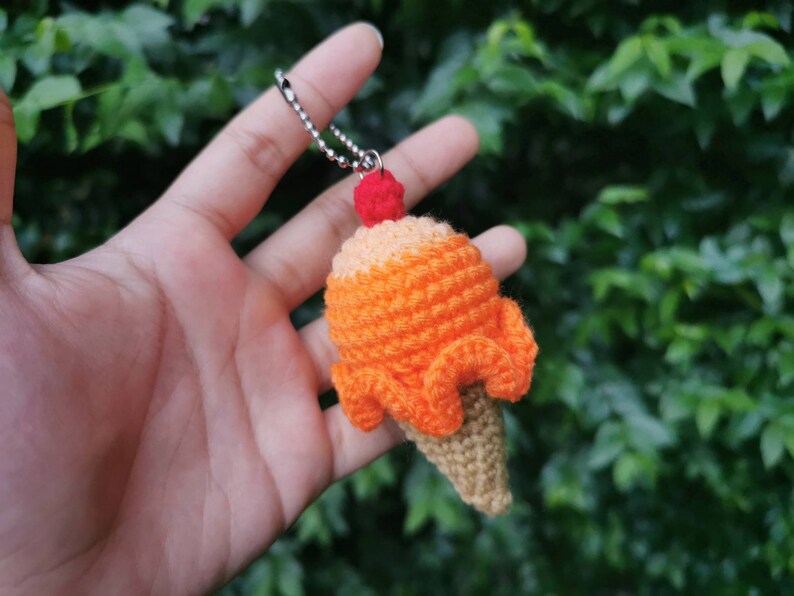 May include: A crocheted ice cream cone keychain with a red cherry on top. The cone is orange and brown and has a silver chain attached.