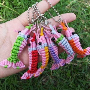 May include: A set of six colorful, crocheted shrimp keychains. Each keychain is made with different colored yarn and features a pink body, black eyes, and a silver chain.