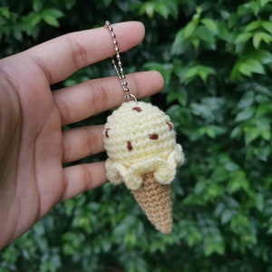 May include: A crocheted ice cream cone keychain with a yellow ice cream scoop and brown chocolate chips. The keychain is attached to a silver chain.