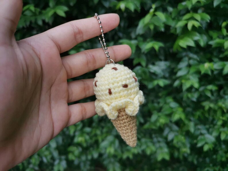 May include: A crocheted ice cream cone keychain with a yellow ice cream scoop and brown chocolate chips. The keychain is attached to a silver chain.