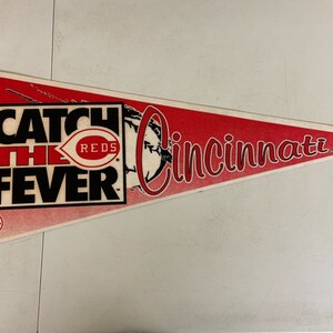 May include: A red and white pennant with the text "Catch the Fever" in black lettering. The Cincinnati Reds logo is featured in the center of the pennant. The text "Cincinnati" is written in a cursive font in the upper right corner of the pennant.