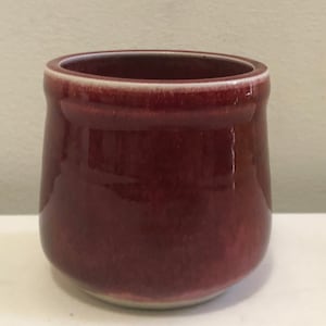 May include: A small, deep red ceramic cup with a white rim. The cup has a smooth, glossy finish.