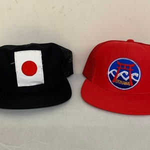 May include: Two black and red mesh trucker hats. The black hat has a white patch with a red circle in the center. The red hat has a blue circle with a red torii gate and waves, and the text "Okinawa" in yellow.
