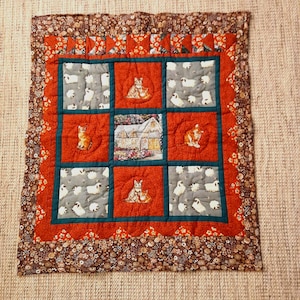 Könnte beinhalten: Handgefertigte Patchwork-Quilt mit quadratischem Design in Rot, Grau und Türkis. Die Quilt zeigt Bilder von Katzen, Schafen und einem Haus. Der Rand ist ein Blumenmuster in Braun- und Rottönen.