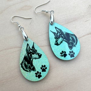 May include: A pair of teal blue teardrop-shaped earrings with a black and white image of a Doberman Pinscher and three black paw prints on each earring.